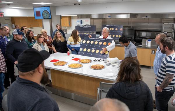 Photo of group surrounding a table with presenter in lab coat showing display board of cheese melt on pizza.