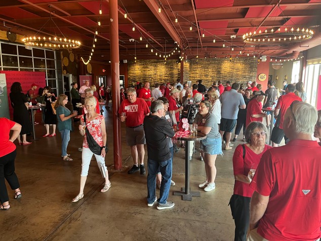 A group largely sporting Wisconsin red mills stands in the Badger State Brewery, talking and enjoying a party.