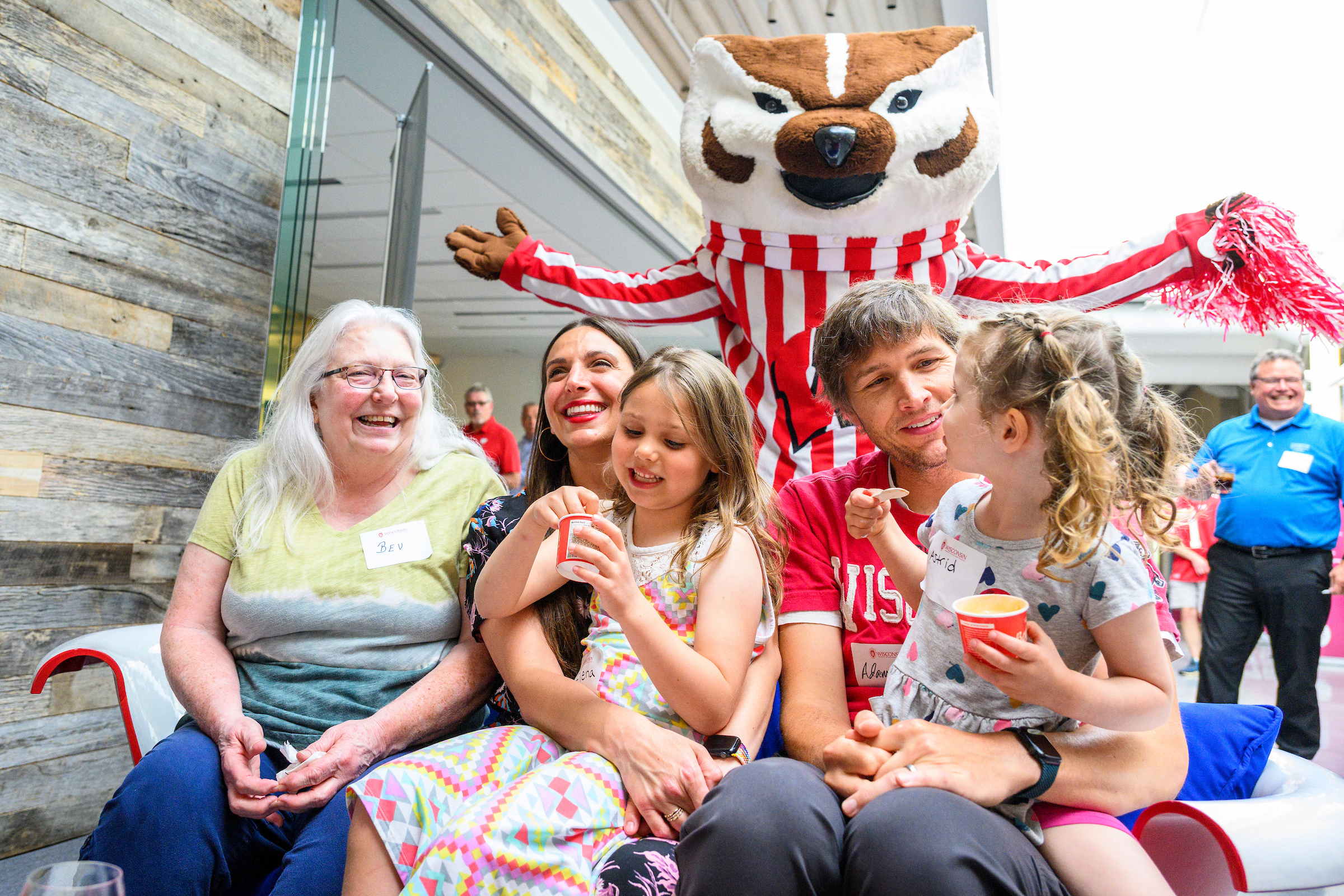 Three adults and two children sit on a bench with Babcock ice cream and pose for a photo with Bucky standing and holding a pompom.