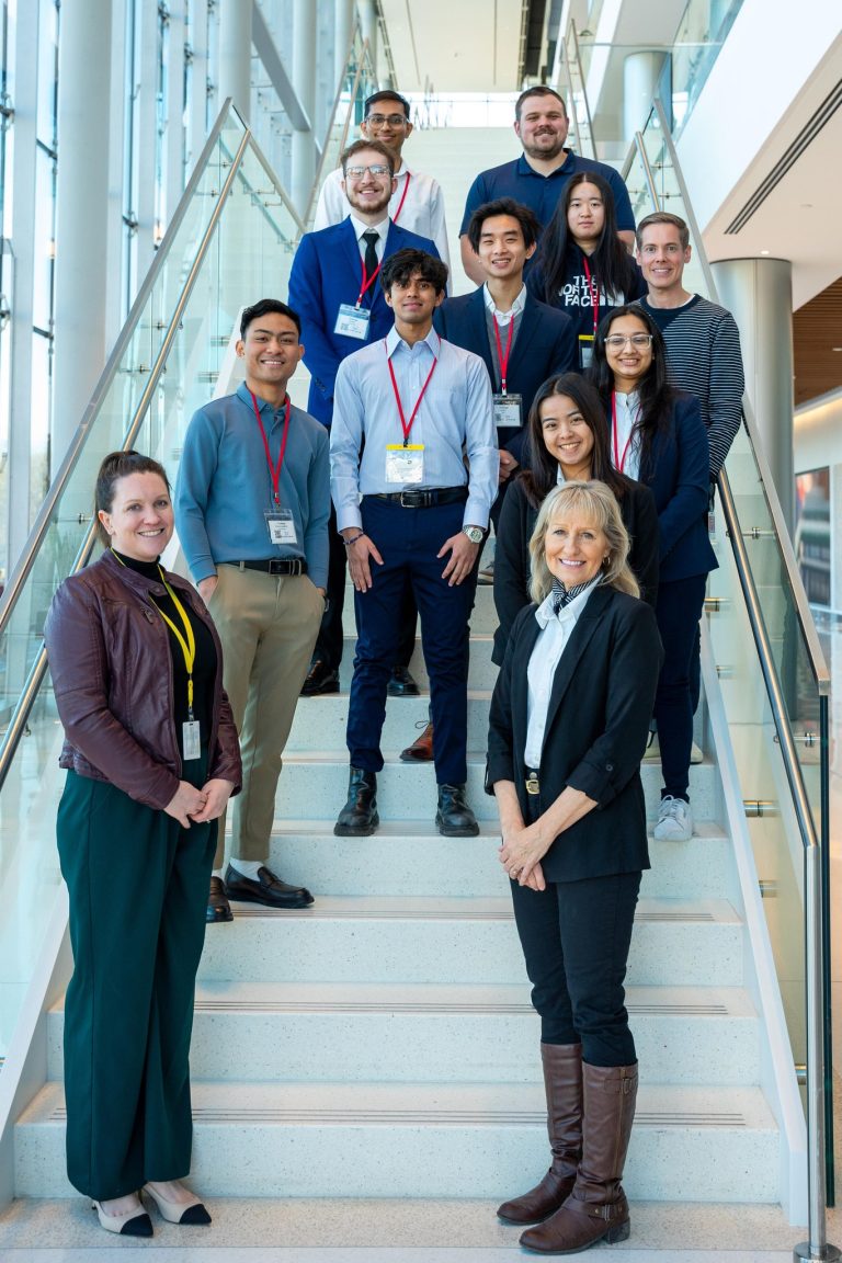 A group of ten people standing on a wide, white staircase inside a modern building with glass walls and bright natural light. Most individuals are wearing business casual attire with name badges and lanyards. Two people are positioned at the bottom of the stairs, while the rest are arranged in rows ascending the steps.