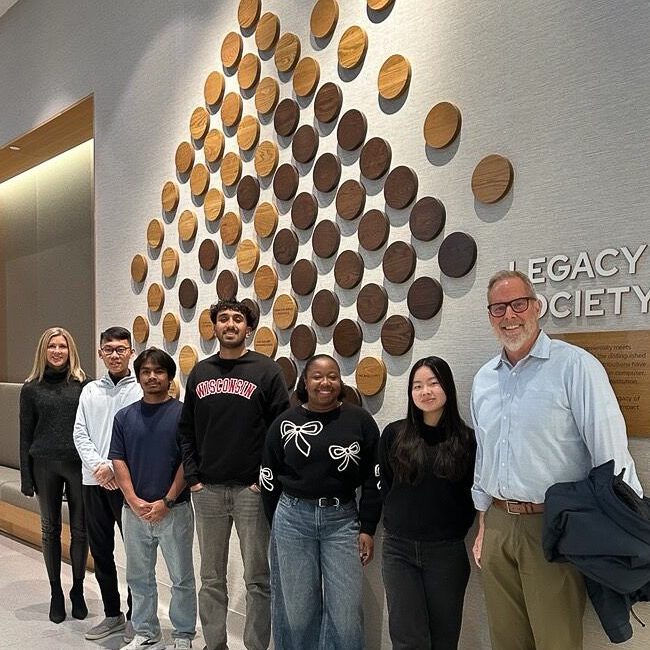 A group of six people standing in a modern hallway in front of a wall display featuring a large arrangement of circular wooden plaques forming a triangular pattern. The wall includes text reading “Legacy Society.” The individuals are dressed in casual and business-casual attire.