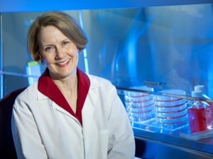 Scientist lynn allen-hoffman smiling facing camera while seated infront of petridishes in a labratory setting