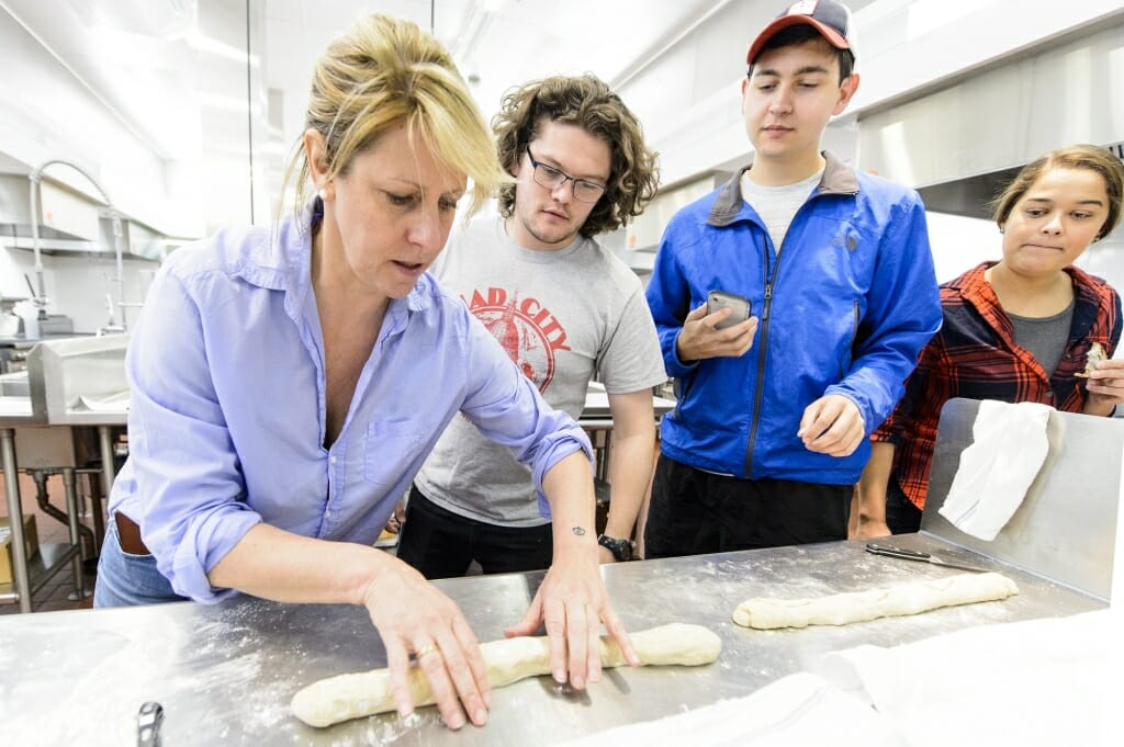 Woman rolling loaves of bread while two males and a female watch on