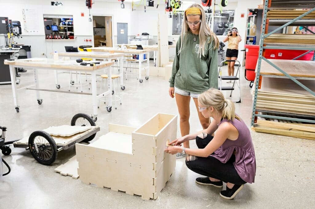 Two women one standing one crouching over a oden box in a technical lab