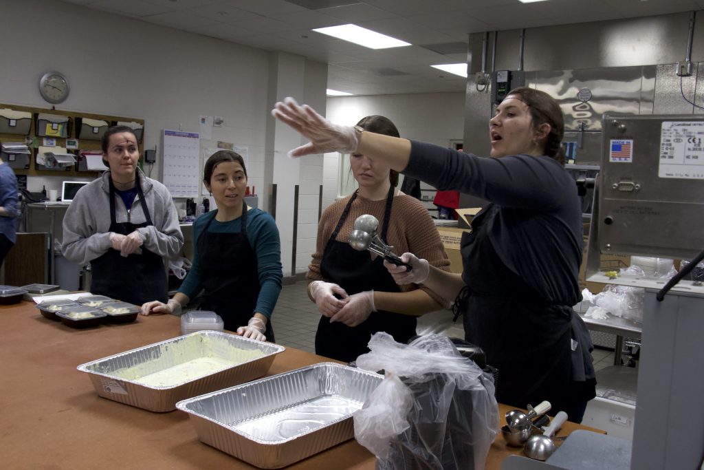 Group stands around food containers and listens to a woman speak. Featured in an industrial kitchen