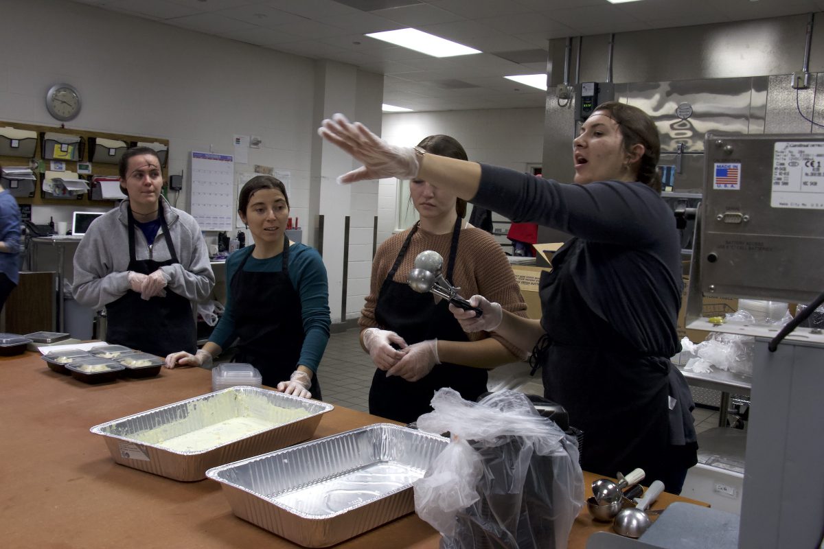 Group stands around food containers and listens to a woman speak. Featured in an industrial kitchen