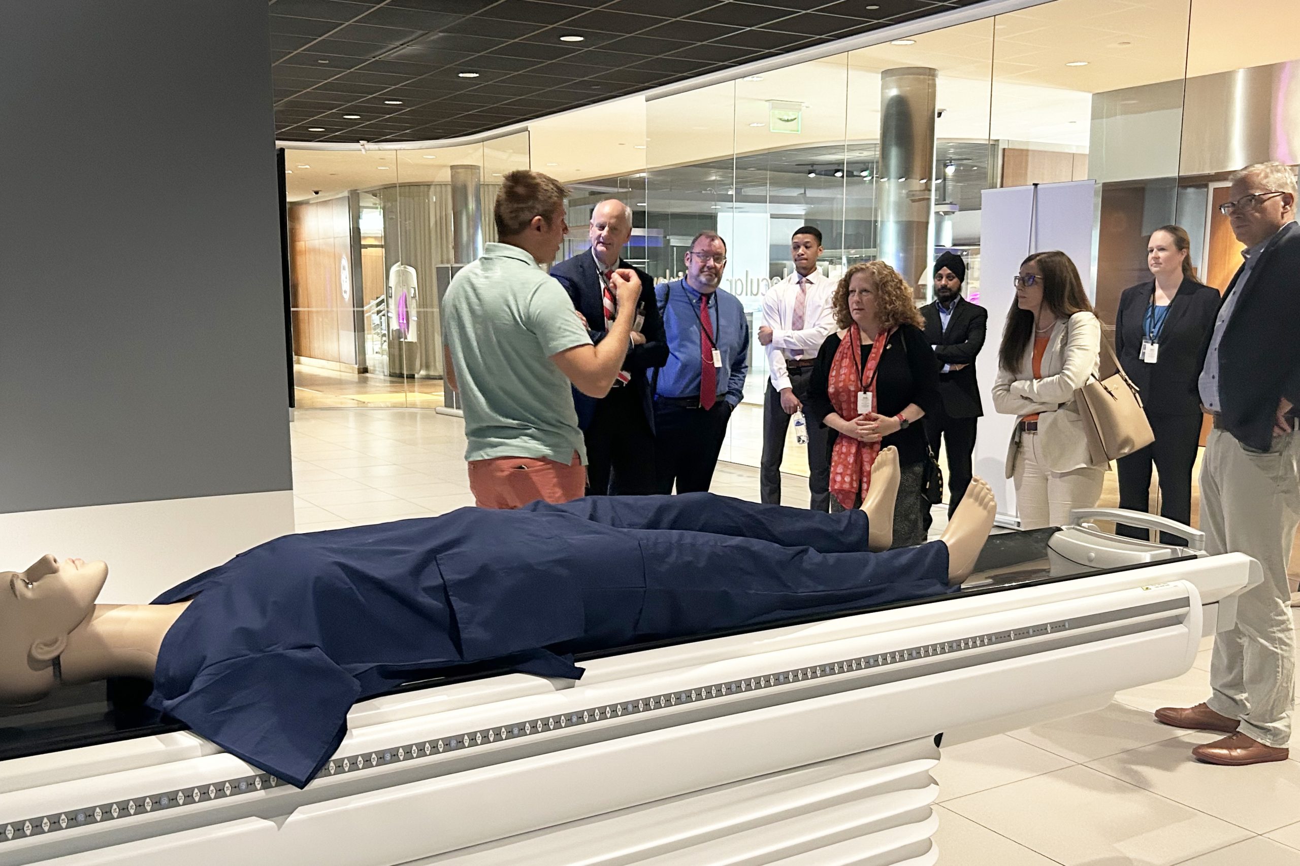 A mannequin in scrubs lies on a table in front of a group dressed in business casual attire. An expert stands in front of the group, appearing to explain the technology that the mannequin is being used to demonstrate.