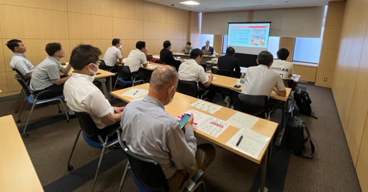 A classroom setting pictured from the back of the room. You can see attendees seated at tables in rows, with papers and notepads out on the table in front of them. At the front of the room facing the camera, John Garnetti is seated next to another person. To his left is a screen with a presentation slide feature U W Madison information.