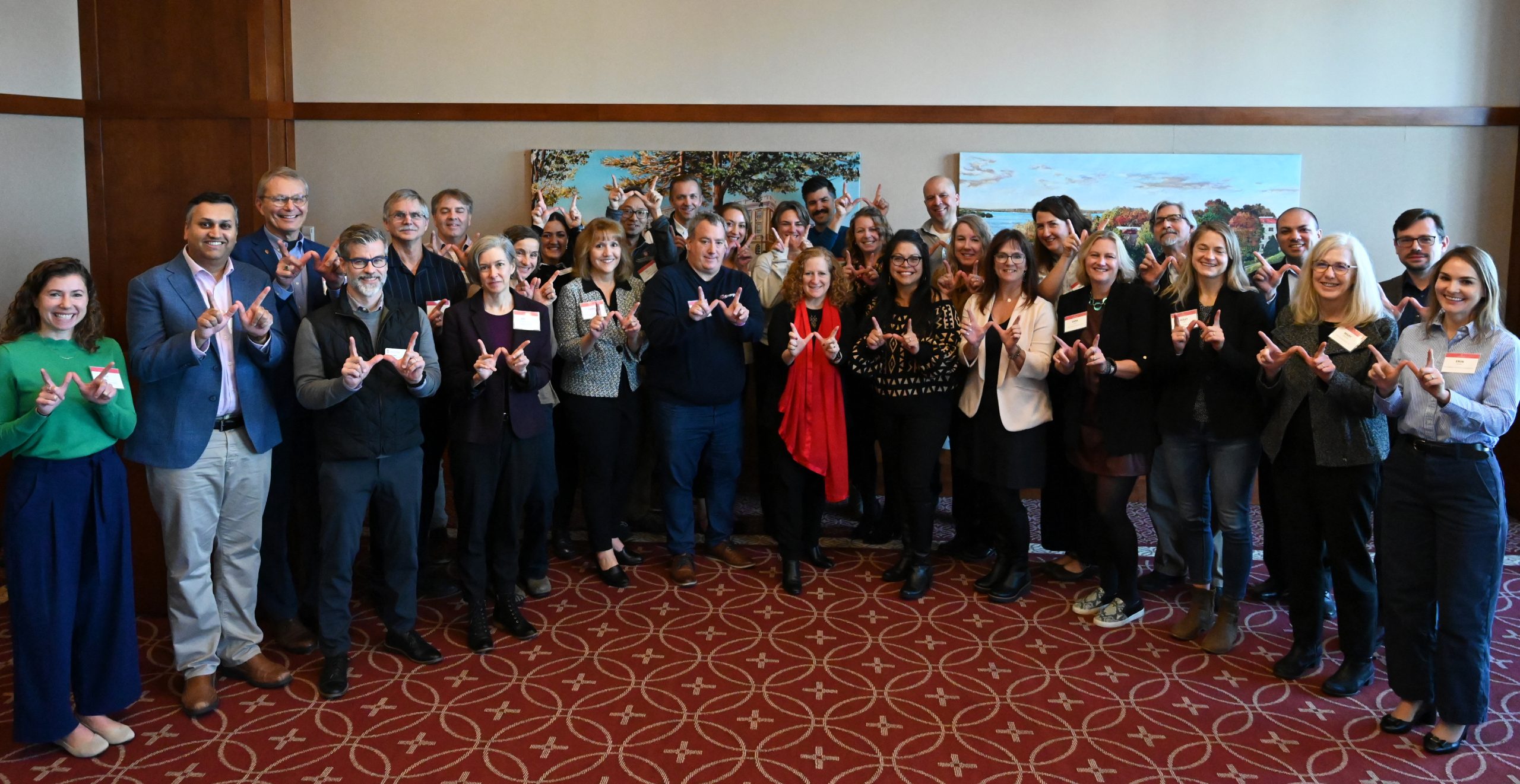 A group of people stand in a semi-circle in a large room, posing for a photo and smiling into the camera. They hold up their hands to each make a "W" gesture.