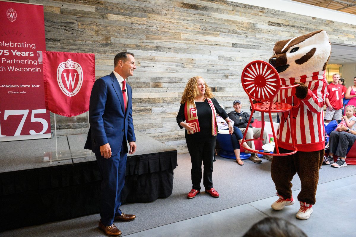 David Kohler stands to the right of Chancellor Mnookin as she gestures toward Bucky Badger holding a red starbust designed chair. Bucky is bringing the chair to David Kohler.