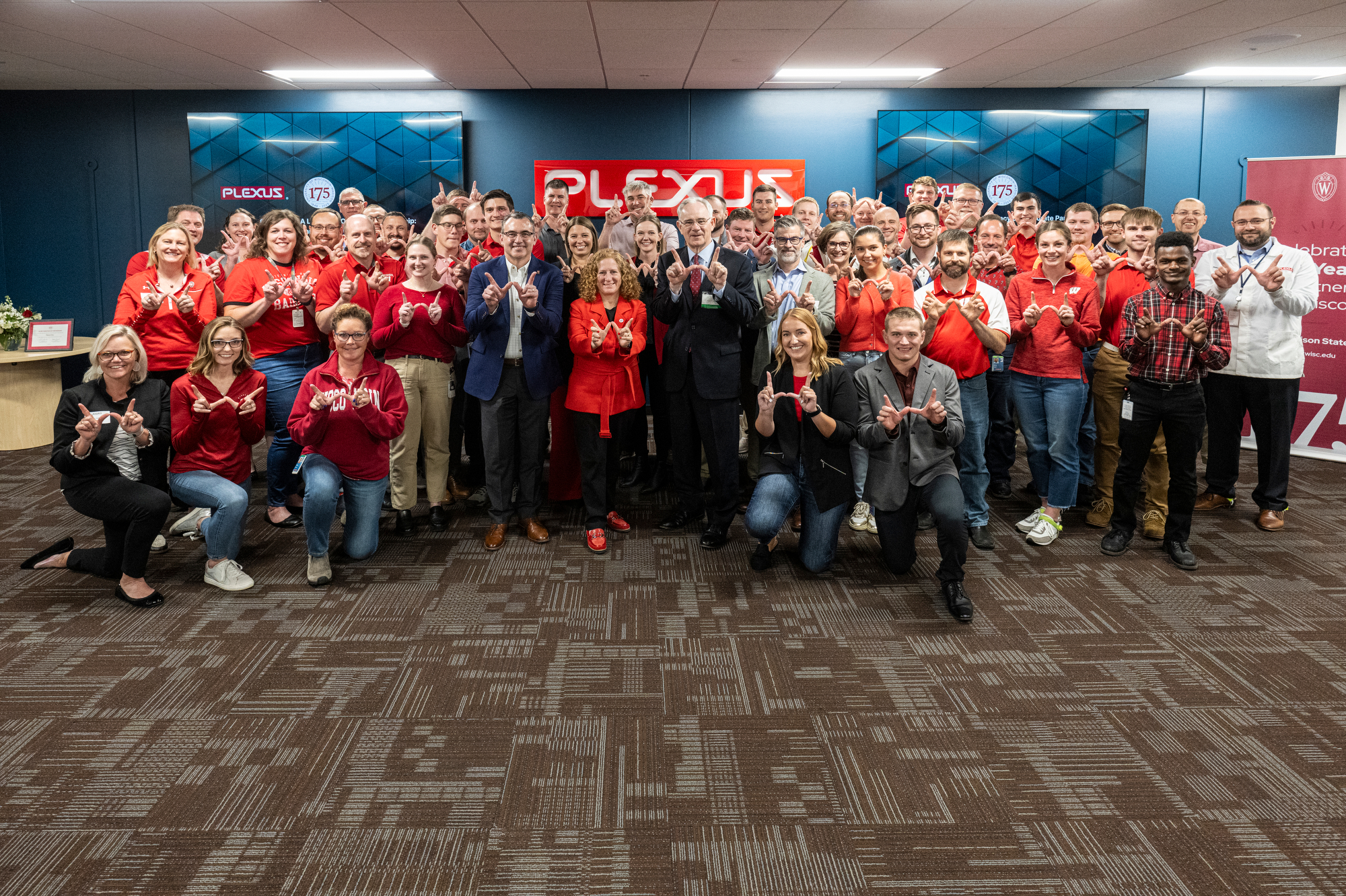 Group of individuals wearing red and grey attire posing with matching u0022Wu0022 hand gestures in an office environment with a u0022Plexusu0022 logo in the background.