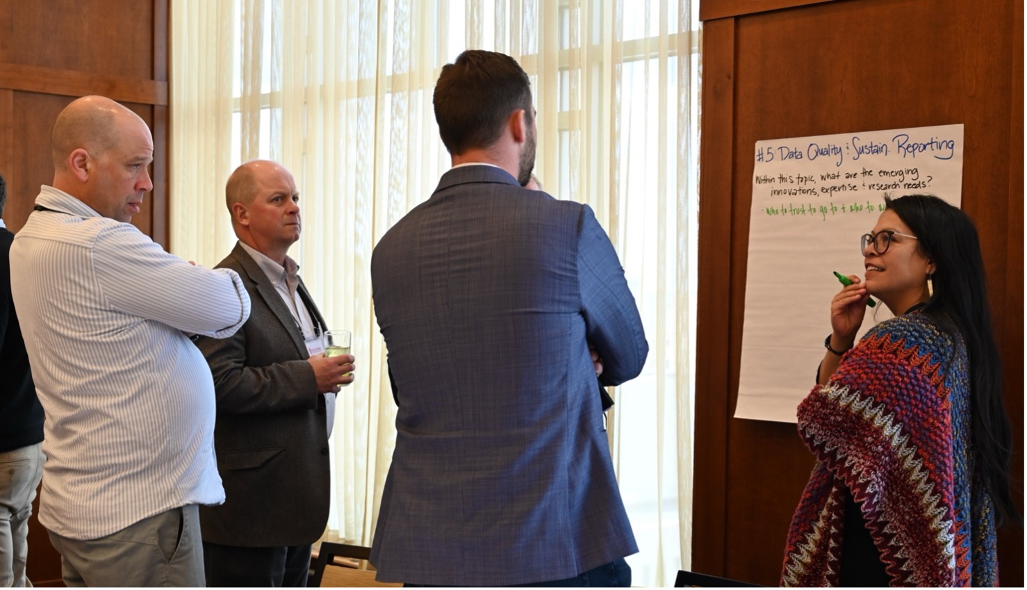 Four professionals are engaged in a discussion near a flip chart in a well-lit conference room with large windows and a wooden wall panel.