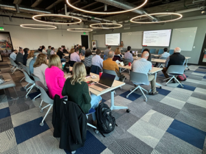 Professional presentation in a conference room with attendees seated facing two speakers, PowerPoint slides displayed in the background.