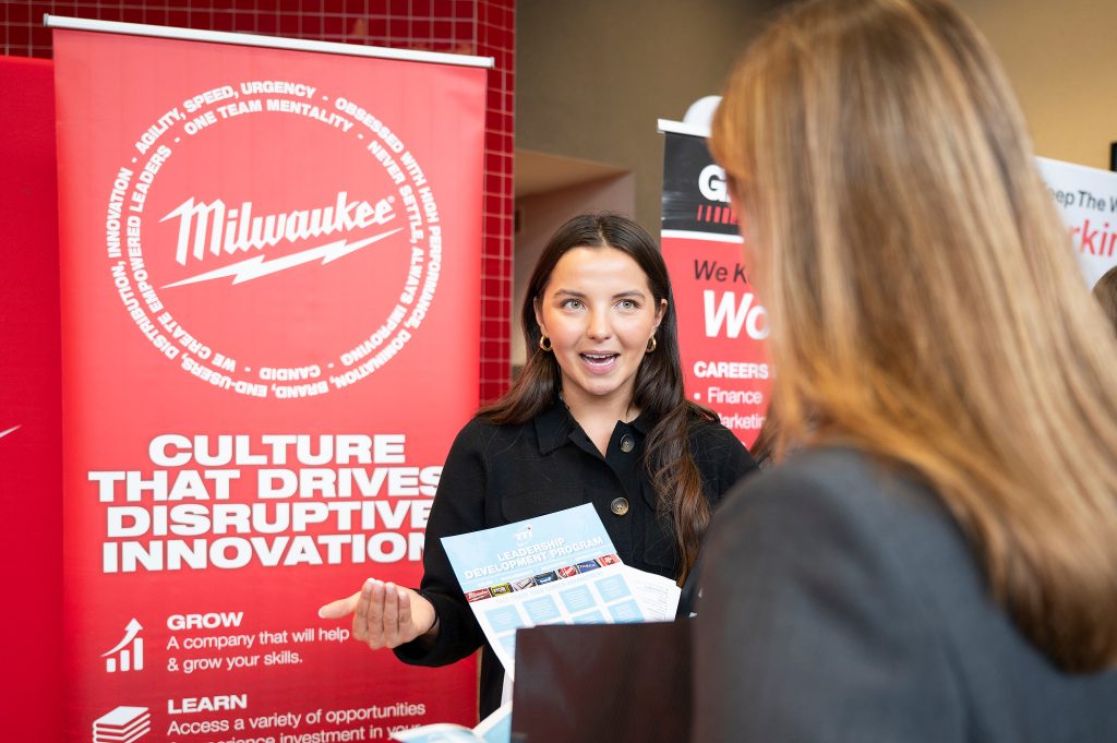 A recruiter from Milwaukee Tool talks to a student. A red sign behind her features the Milwaukee logo with white text under that reads "Culture that drives disruptive innovation."