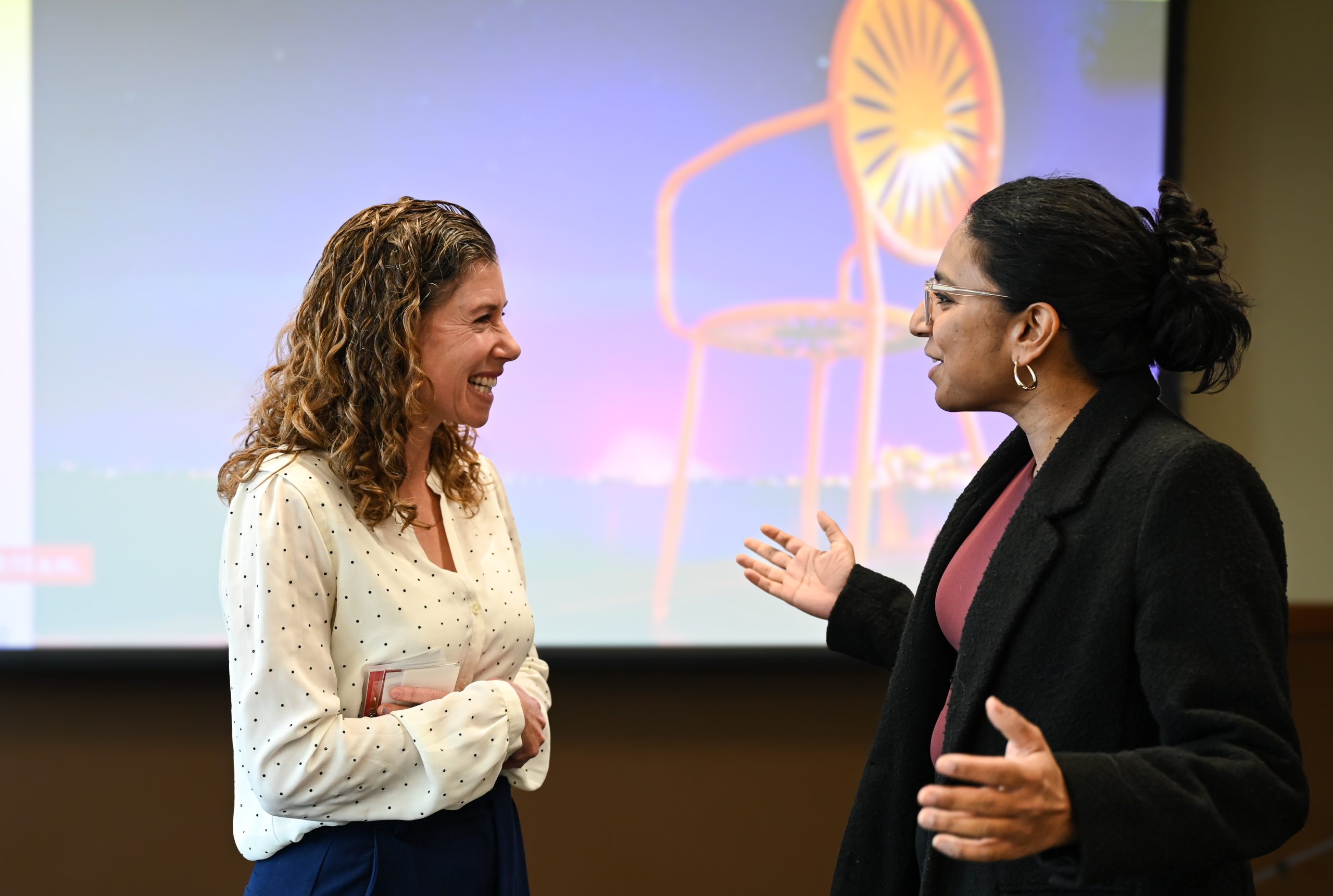 Two people converse in front of a screen with an orange terrace chair.