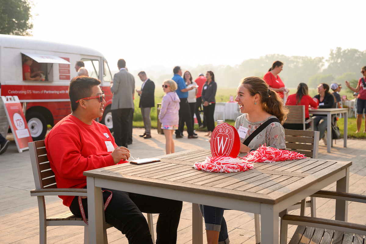 Two alumni are sitting at a wooden table outdoors and having a conversation.