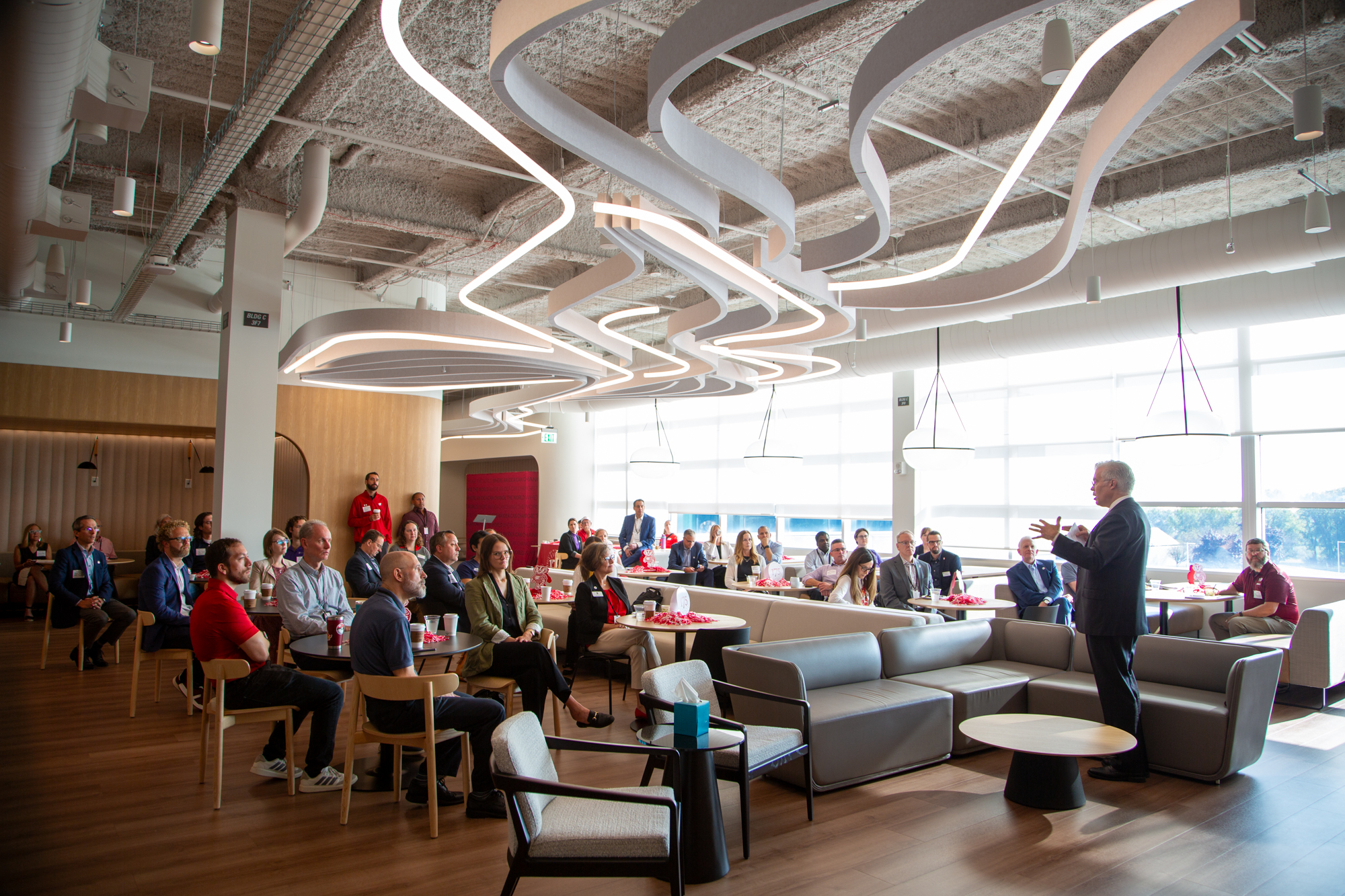 Approximately 40 people sit at tables and lounge seating in a modern event space while a speaker addresses the audience near large windows.