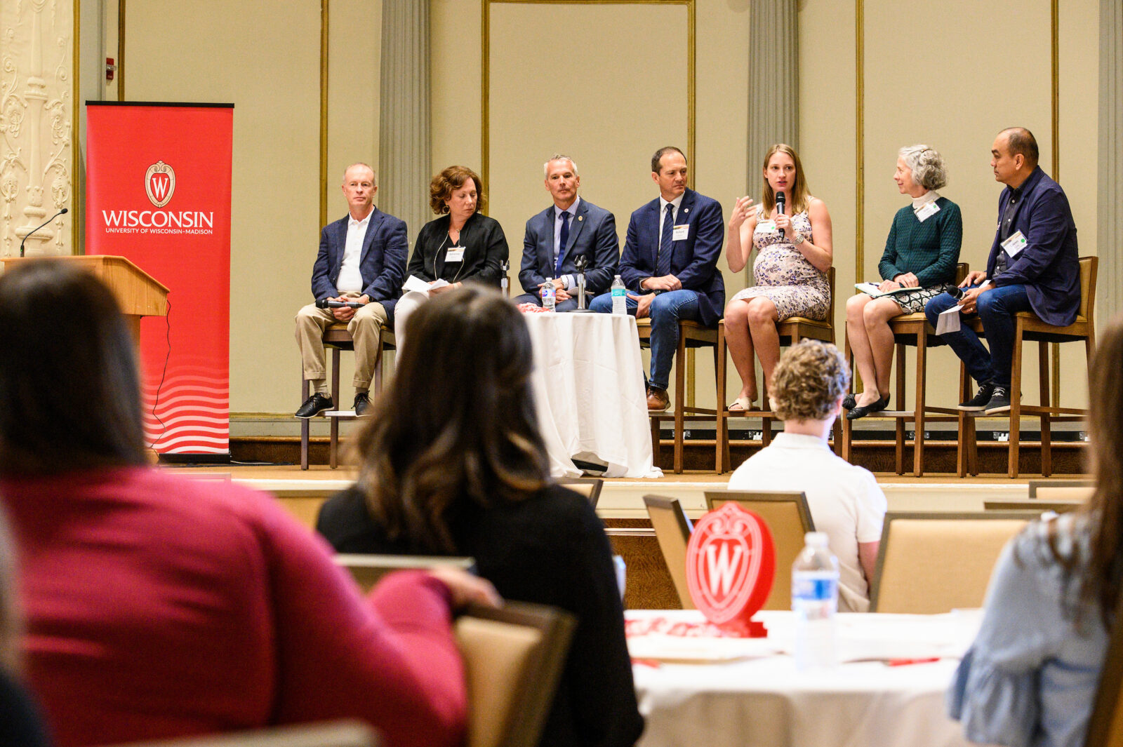 A panel of seven people sit on chairs on a stage. One of them speaks into a microphone. UW branding is visible on a banner and on a table.