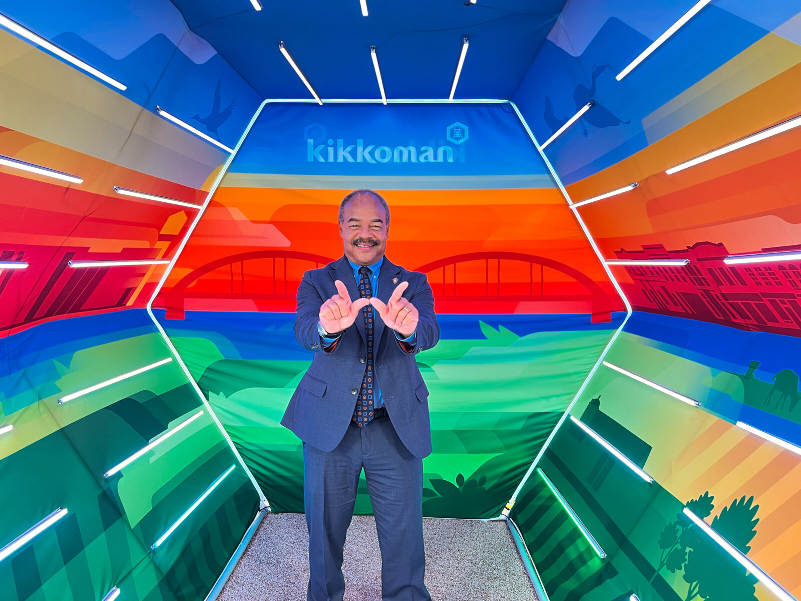 Eric Wilcots stands in a colorful Kikkoman display, holding his hands in the shape of a “W,” with the Kikkoman logo above him.